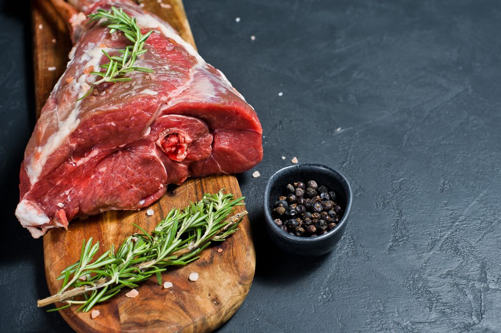 Raw leg of goat on a wooden cutting Board. Rosemary, thyme, black pepper. Black background, side view, space for text