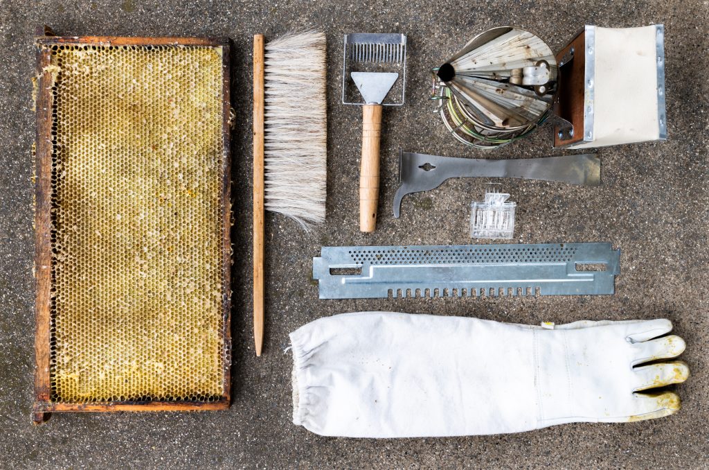 Overhead view of beekeeping equipment including a smoker, frame, glove, hive tool, queen clip, mouse guard, and uncapping tool