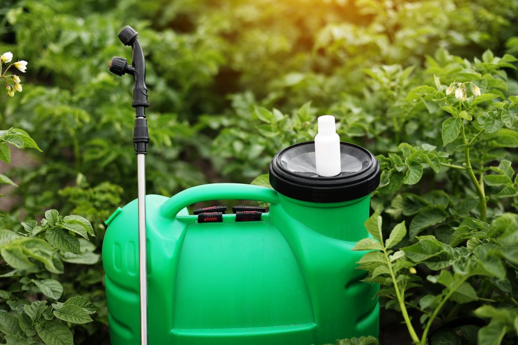White little bottle of pesticide, herbicide for protecting plants from diseases and pests with mock up stands on container sprayer on natural green garden background. Agricultural seasonal work.