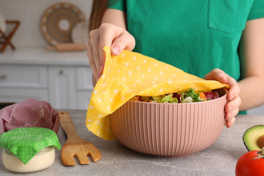 Woman covering bowl of fresh salad with beeswax food wrap at light grey table in kitchen, closeup