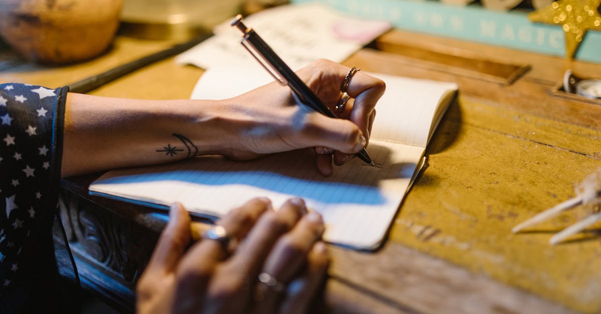 a hand with tattoos writing in a notebook on a rustic wooden desk under warm lighting
