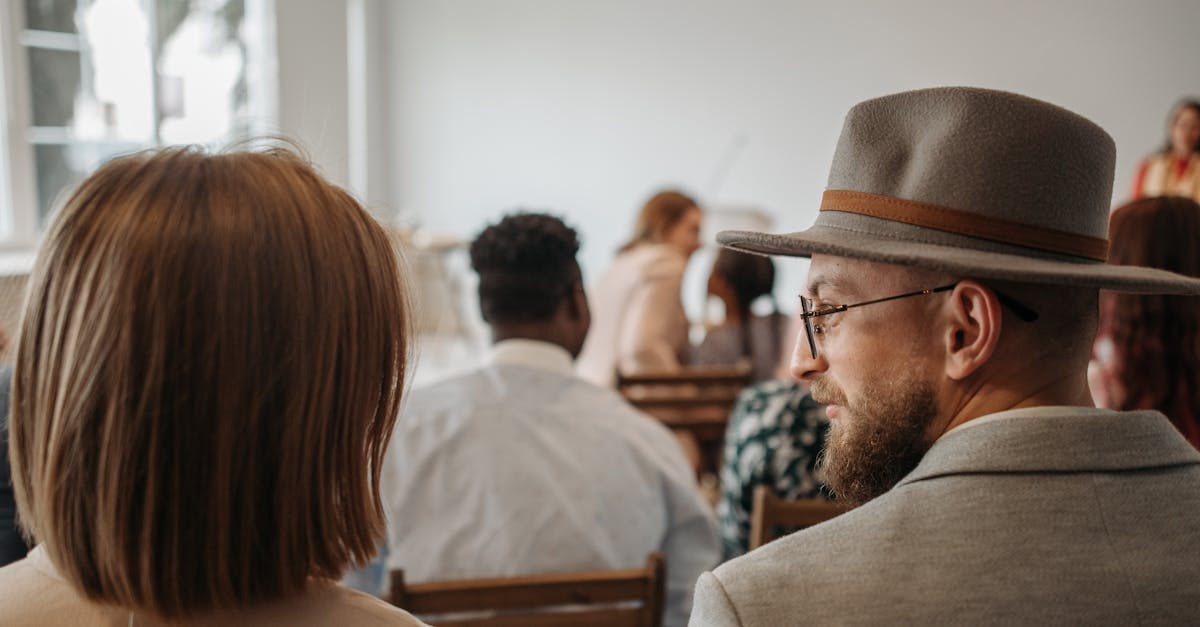 a man wearing a hat and glasses sits among attendees at an indoor seminar 1