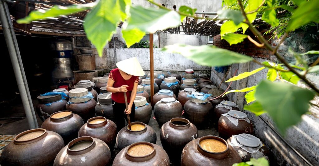 a person in a conical hat checks large fermentation jars in a sunny outdoor courtyard