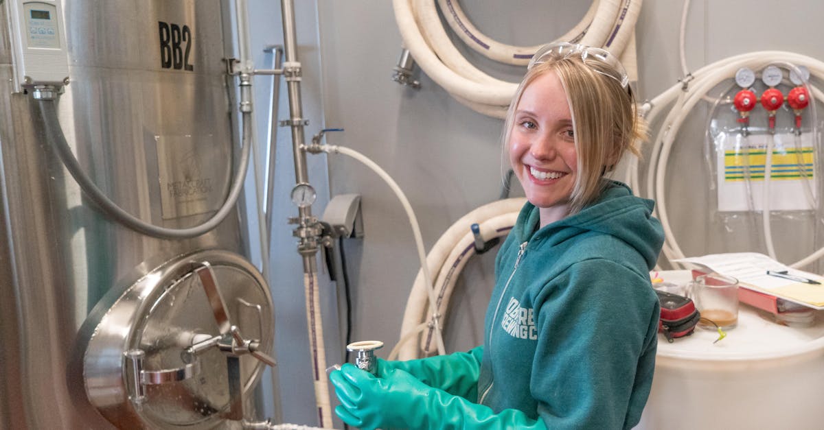 a woman in a green jacket and gloves working in a brewing factory smiling near stainless steel equi