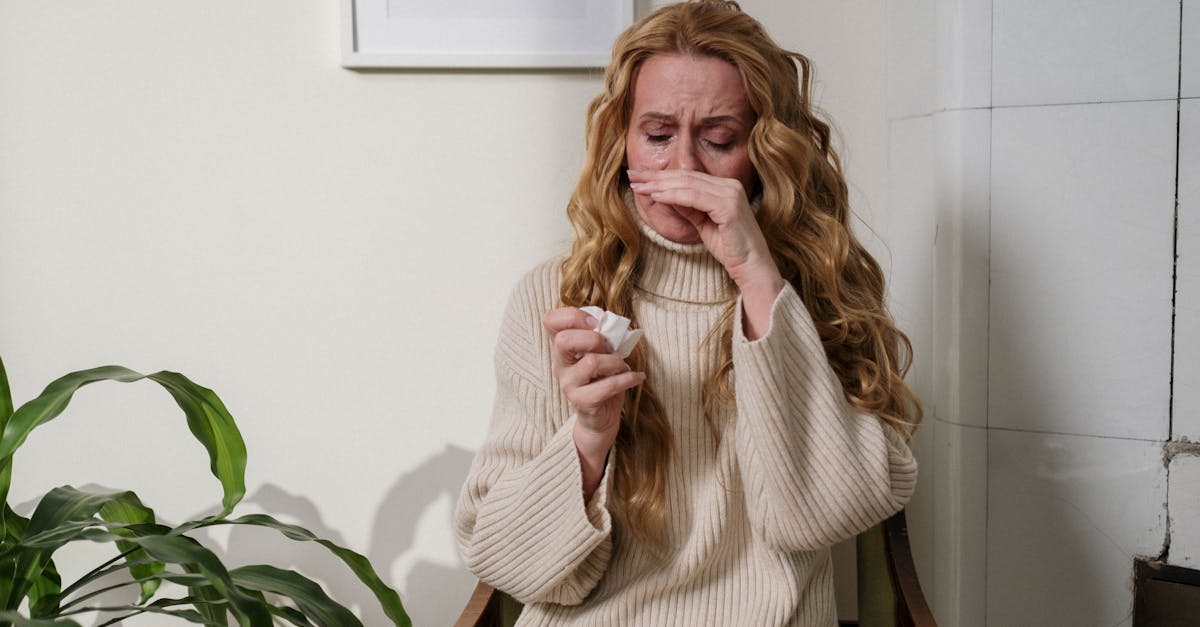 a woman with a runny nose sitting indoors using a tissue to relieve her allergies