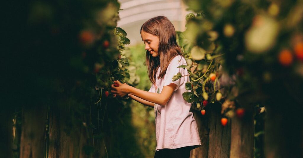 a young girl happily picks strawberries in an indoor greenhouse surrounded by lush plants