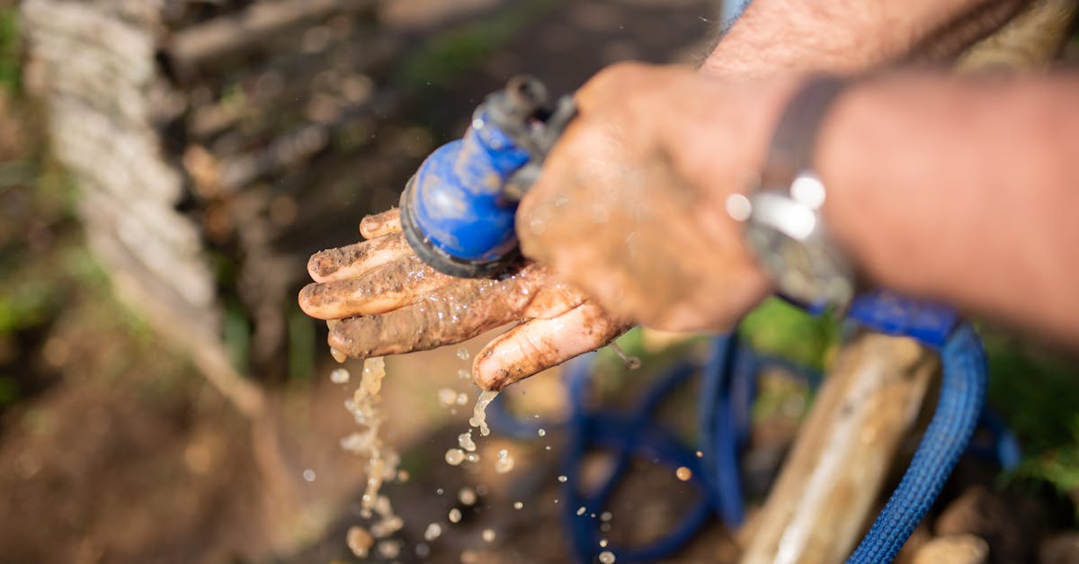 close up of muddy hands being cleaned with a blue garden hose splashing water outdoors 1