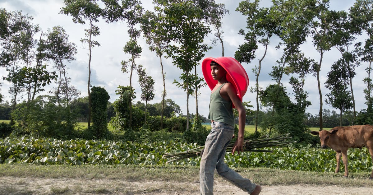 young boy carrying a bright red basket on his head in a lush rural area during the day