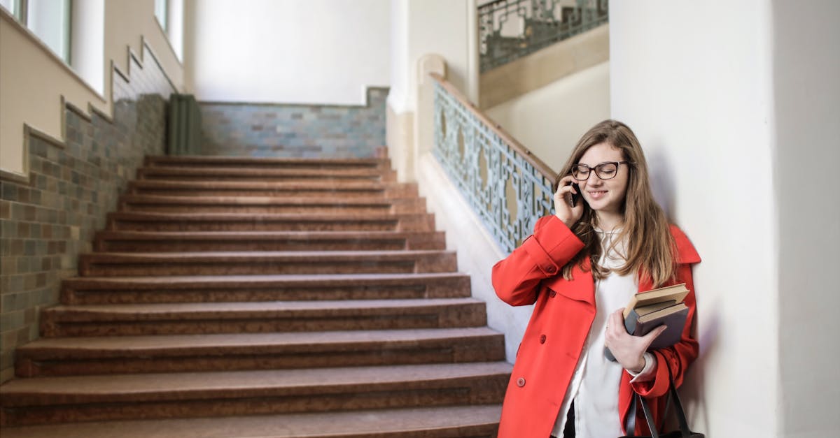 young woman in red coat smiling while using a smartphone indoors holding books