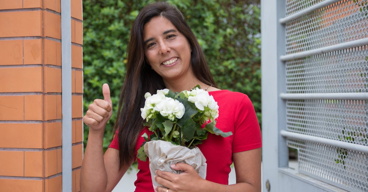 a smiling woman in a red shirt holds a white flower bouquet standing by a gate outdoors