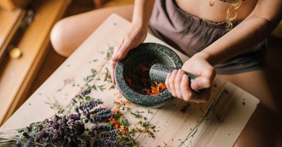 close up of hands using mortar and pestle to grind herbs surrounded by dried flowers on wooden surf