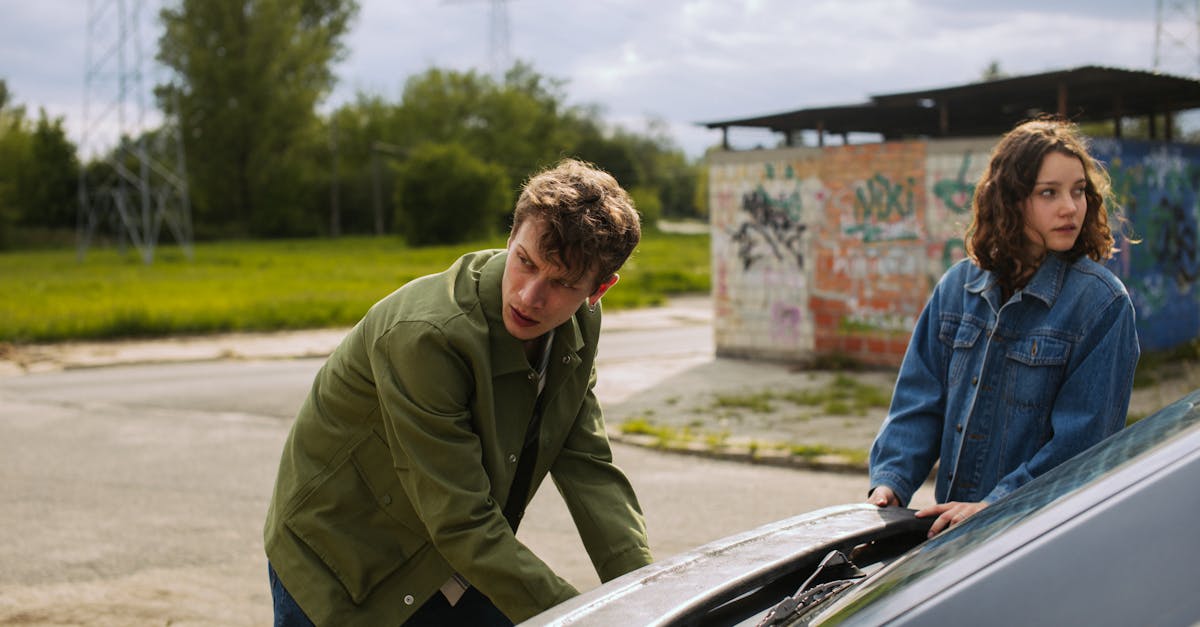 two young adults examining a car engine in an urban area surrounded by graffiti