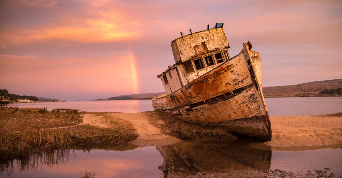 a rusted abandoned ship rests ashore under a serene sunset sky with a distant rainbow