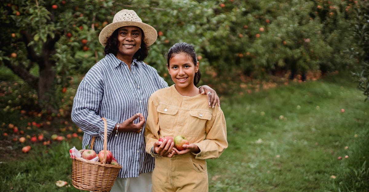 content ethnic female harvester with wicker basket embracing charming girl on path near apple trees