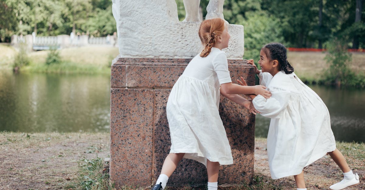 two young girls in white dresses playing near a statue in a scenic park